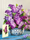 A large, paper bouquet of lilacs in a blue and white pattern vessel, with a matching card standing next to it on a blue wooden table. 