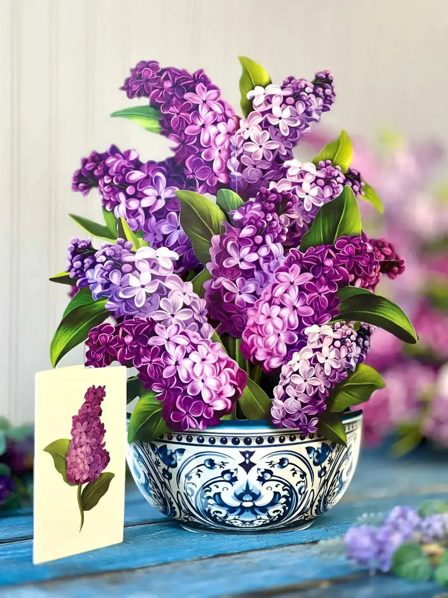 A large, paper bouquet of lilacs in a blue and white pattern vessel, with a matching card standing next to it on a blue wooden table. 