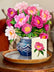 A paper bouquet of pink peonies in a blue and white patterned vase, sitting atop a wooden table with a matching card next to it. 