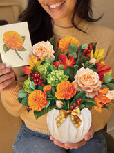 A smiling woman with the top half of her face cut out of frame holds a large paper bouquet made of peach, orange and green flowers, red berries, and leaves in her left hand, and a matching card in her right hand. 