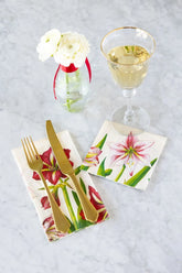 A guest napkin and cocktail napkin printed with white and red amaryllis flowers on a white marble background. A set of gold silverware rests on the guest napkin to the left, a champagne glass sits atop the cocktail napkin, and a bud vase with two white ranunculus sits at the top left of the photo.