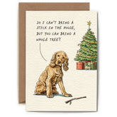 Greeting card of a yellow dog with a stick laying next to it. A Christmas tree can be seen in the background. The dog has a speech bubble above it that says "So I can't bring a stick in the house, but you can bring a whole tree?"