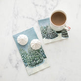 A guest napkin and cocktail napkin printed with an illustration of evergreen trees covered in snow sit on a white marble background. Two meringue  puffs and a mug with hot chocolate sit on top of the napkins.