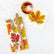 A guest napkin and cocktail napkin printed with orange, yellow and red fall leaves sit on a white marble background. A set of gold silverware rests on the guest napkin and a glass with an amber colored drink sits atop the cocktail napkin.