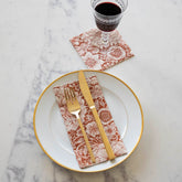 A white dinner plate with gold trim sits on a white marble background, dressed with a guest napkin and gold silverware. A cocktail napkin with a goblet of wine sits to the top right of the photo. Both napkins have a burnt orange and white floral pattern.