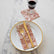 A white dinner plate with gold trim sits on a white marble background, dressed with a guest napkin and gold silverware. A cocktail napkin with a goblet of wine sits to the top right of the photo. Both napkins have a burnt orange and white floral pattern.