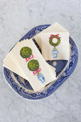 A large blue chinoiserie dish sits on a white marble background. Inside of the dish are a stack of guest napkins and a set of cocktail napkins, each printed with illustrations of a holiday topiary.