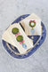 A large blue chinoiserie dish sits on a white marble background. Inside of the dish are a stack of guest napkins and a set of cocktail napkins, each printed with illustrations of a holiday topiary.