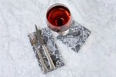 A guest napkin and cocktail napkin printed with a black and white illustration of ravens, flowers and spiderwebs sit on a white marble background. A set of silverware rests on the guest napkin and a wine glass sits atop the cocktail napkin.