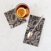 A cocktail napkin and guest napkin printed with brown and white sunflower illustrations on a white marble background. A whiskey glass sits on the cocktail napkin, and a silver spoon rests on the guest napkin.