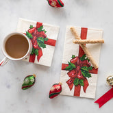 A cocktail napkin and guest napkin with an illustration of a red ribbon tied with holly leaves sit on a white marble background. The napkins are styles with small Christmas bulbs, wafer cookies, and a mug of hot chocolate.