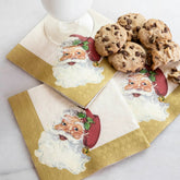 A stack of three cocktail napkins with gold trim and a vintage Santa Claus, underneath five chocolate chip cookies and a glass of milk, laid on a white background.