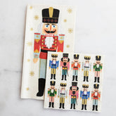 A guest napkin with a nutcracker dressed in red, grey and gold, laid next to a cocktail napkin with a repeating print of nutcrackers dressed in varying colors. The two napkins are laid on a white marble background.
