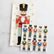 A guest napkin with a nutcracker dressed in red, grey and gold, laid next to a cocktail napkin with a repeating print of nutcrackers dressed in varying colors. The two napkins are laid on a white marble background.