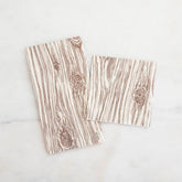 A guest napkin and cocktail napkin both printed with a white and brown wood pattern lay next to each other on a white marble background.