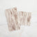 A guest napkin and cocktail napkin both printed with a white and brown wood pattern lay next to each other on a white marble background.