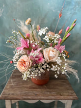 Floral arrangement made in pink and peach tones on a babies breath base, in a clay vase. Displayed on a wooden table in front of a blue-grey background.