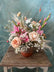 Floral arrangement made in pink and peach tones on a babies breath base, in a clay vase. Displayed on a wooden table in front of a blue-grey background.