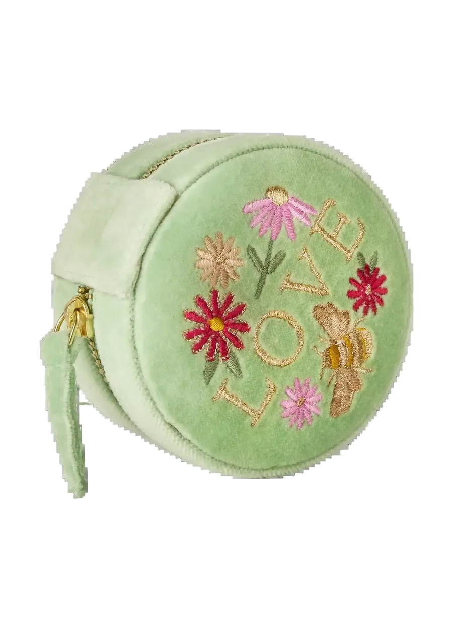 A light green velvet round jewelry box with embroidered flowers, a bee, and the word 'LOVE' on the front, featuring a side strap and a gold zipper.
