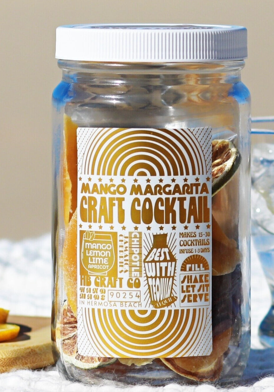 A clear glass jar filled with dried botanicals and citrus slices, sealed with a white lid, and labeled 'Mango Margarita Craft Cocktail' and 'HB Craft Co.'. The jar is photographed outdoors on a light surface with a softly blurred neutral background.