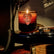 A brown and amber colored jar candle with a guitar pick emblem on the front sits on the bottom end of a fender guitar, which is mostly out of frame. A fender amp and other musical elements can be seen out of focus in the background.