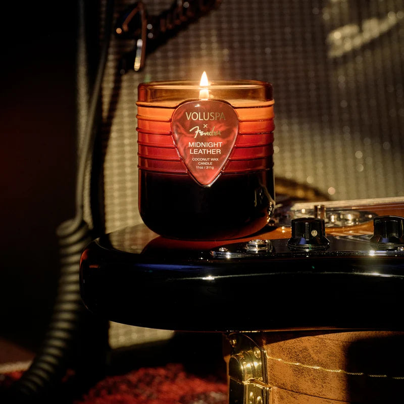 A brown and amber colored jar candle with a guitar pick emblem on the front sits on the bottom end of a fender guitar, which is mostly out of frame. A fender amp and other musical elements can be seen out of focus in the background.