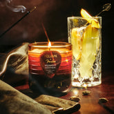 A brown and amber colored jar candle with a guitar pick emblem on the front sits on a dark leather setting, with cardamom pods, a leather jacket, cocktail glass, and burning incense placed around it.