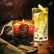 A brown and amber colored jar candle with a guitar pick emblem on the front sits on a dark leather setting, with cardamom pods, a leather jacket, cocktail glass, and burning incense placed around it.