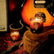 A warm amber tones stylized photo with a fender guitar and amp partially in frame sit behind two candles in glass jars. One candle sits atop a brown velvet guitar case, while the other is on the floor.