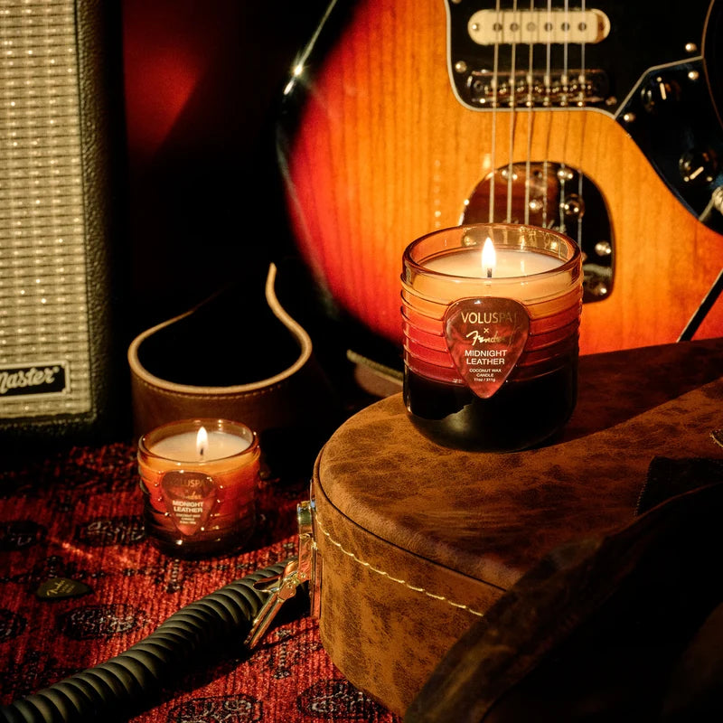 A warm amber tones stylized photo with a fender guitar and amp partially in frame sit behind two candles in glass jars. One candle sits atop a brown velvet guitar case, while the other is on the floor.
