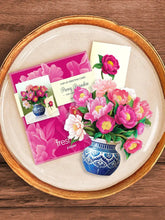 Pop-up card of pink peonies in a blue and white vase, laid on top of the card's envelope, and a matching greeting card. The items are displayed as a flat-lay photo, on top of a ceramic plate on a light wood table.