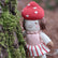 Hand-knit doll with a mushroom hat and red and white striped skirt sitting next to a mossy tree.