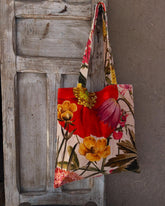 A velvet tote bag with a large orange, red, and pink floral print design hangs on the handle of a grey-washed wooden door.