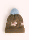 A light brown children's knit hat with orange highland cows, white westies, blue polka dots, and a powder blue pompom on the top, against a white background.