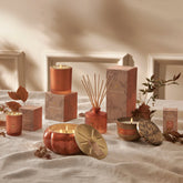 Candle and fragrance diffuser collection with decorative boxes and candles on a textured surface. The candles and diffusers are in warm copper-orange and light taupe colors, set against a beige background. Leaves and other autumnal elements sit around the products.
