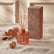 Small fragrance diffuser in a matte copper-orange color, with reeds sitting inside of it, displayed next to it's product box on a tabled with beige fabric. Autumnal elements sit next to the products.