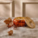Candle in a pumpkin-shaped holder with a leaf and branch on a textured beige surface.
