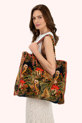 A woman wearing a white sleeveless top and a light-colored knit skirt carries a large quilted tote bag with a detailed woodland print featuring owls, red mushrooms, and green foliage.