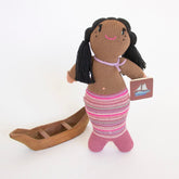 Hand-knit black mermaid doll with a pink striped tail, holding a book and standing next to a boat, against a white background. 