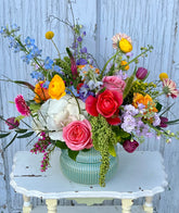 A vibrant, multicolor floral arrangement with pink roses, white kale, gerbera daisies, delphinium, and more in a teal ceramic vessel, sitting on top of a distressed, white table in front of a weathered background.