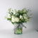 A floral arrangement of white flowers and green foliage in a clear glass jar with a gray ribbon, set against a plain light gray background.