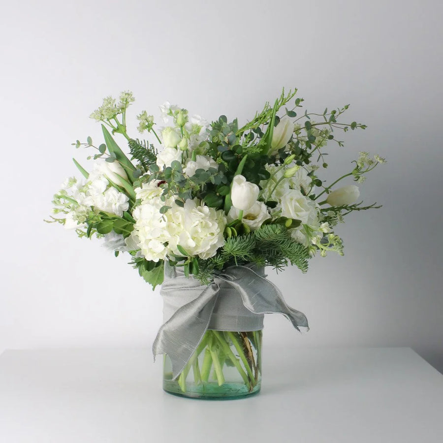 A floral arrangement of white flowers and green foliage in a clear glass jar with a gray ribbon, set against a plain light gray background.