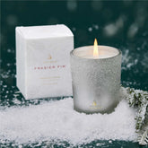 A silver votive candle with white flocking in front of its accompanying box, surrounded by fake snow against a dark forrest green backgroud.
