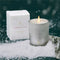 A silver votive candle with white flocking in front of its accompanying box, surrounded by fake snow against a dark forrest green backgroud.