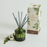 A green glass reed holder with reeds sticking out of it, with the accompanying box posed behind it. A silver Christmas bulb and a sprig of pine sit to the left of the diffuser.