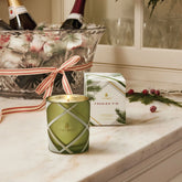 A green and white plaid candle sit in front of its accompanying box, styled with wine bottles in an ice bucket and Christmas greens behind it on a marble counter.