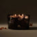 A lit candle in a black and grey smokey glass sits against a grey-brown background, with amber colored crystals around it.