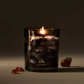 A lit candle in a black and grey smokey glass sits against a grey-brown background, with amber colored crystals around it.