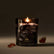 A lit candle in a black and grey smokey glass sits against a grey-brown background, with amber colored crystals around it.