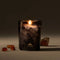 A lit candle in a black and grey smokey glass sits against a grey-brown background, with amber colored crystals around it.